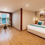 A spacious and luxurious Suite aboard the MS Alma Nile Cruise, featuring a comfortable king-size bed, warm wood flooring, and a large sliding glass door opening to a private balcony overlooking the Nile River.
