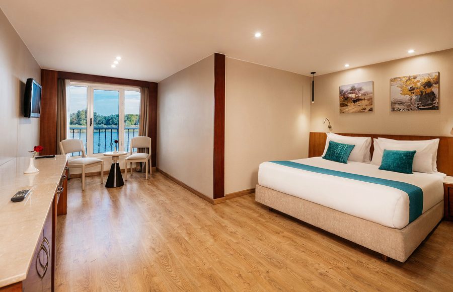 A spacious and luxurious Suite aboard the MS Alma Nile Cruise, featuring a comfortable king-size bed, warm wood flooring, and a large sliding glass door opening to a private balcony overlooking the Nile River.