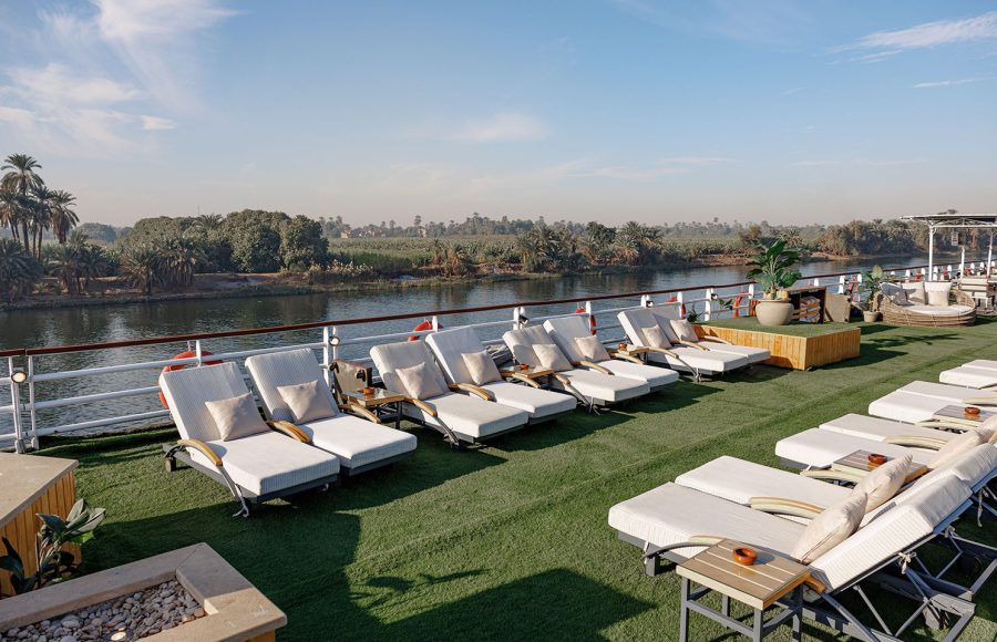 Comfortable lounge chairs and sunbeds lined up on the lush Sun Deck of the MS Alma Nile Cruise, offering panoramic views of the Nile River and green riverbanks.