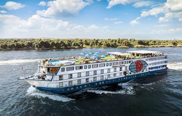 An aerial view of the MS Chateau Lafayette Nile Cruise ship sailing along the Nile River, showcasing its expansive sun deck, swimming pool, and distinct branding.