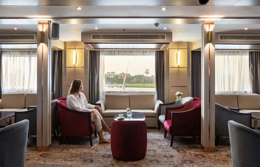 A guest relaxing in the elegant restaurant lounge on the MS Chateau Lafayette Nile Cruise, featuring stylish seating and large panoramic windows overlooking the riverbanks.