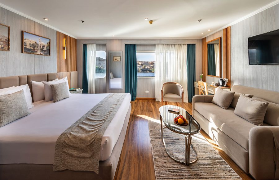 A spacious and luxurious suite aboard the MS Chateau Lafayette Nile Cruise, featuring a plush king-size bed, a comfortable sitting area, and large windows with river views.
