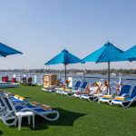 Guests relaxing on loungers and blue umbrellas on the Sun Deck of the MS Chateau Lafayette Nile Cruise, with panoramic views of the Nile River.