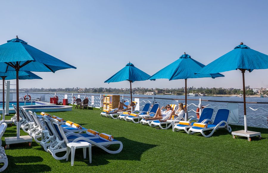 Guests relaxing on loungers and blue umbrellas on the Sun Deck of the MS Chateau Lafayette Nile Cruise, with panoramic views of the Nile River.