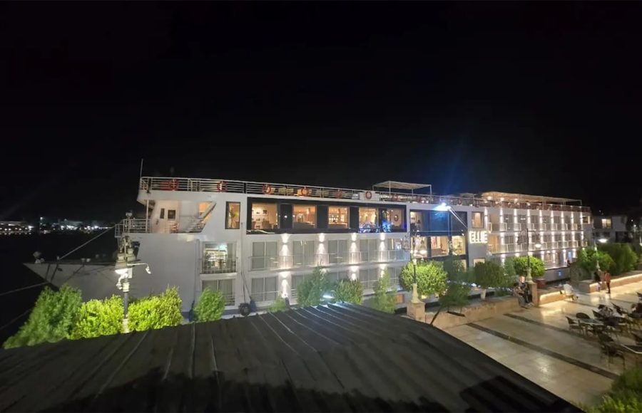 The MS Ellie Nile Cruise ship docked at night, brightly lit with exterior lighting against a dark sky and reflected on the boardwalk.
