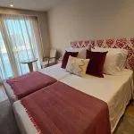 A cruise cabin with two twin beds featuring a red and white patterned headboard, a large window with sheer curtains, and wood flooring.