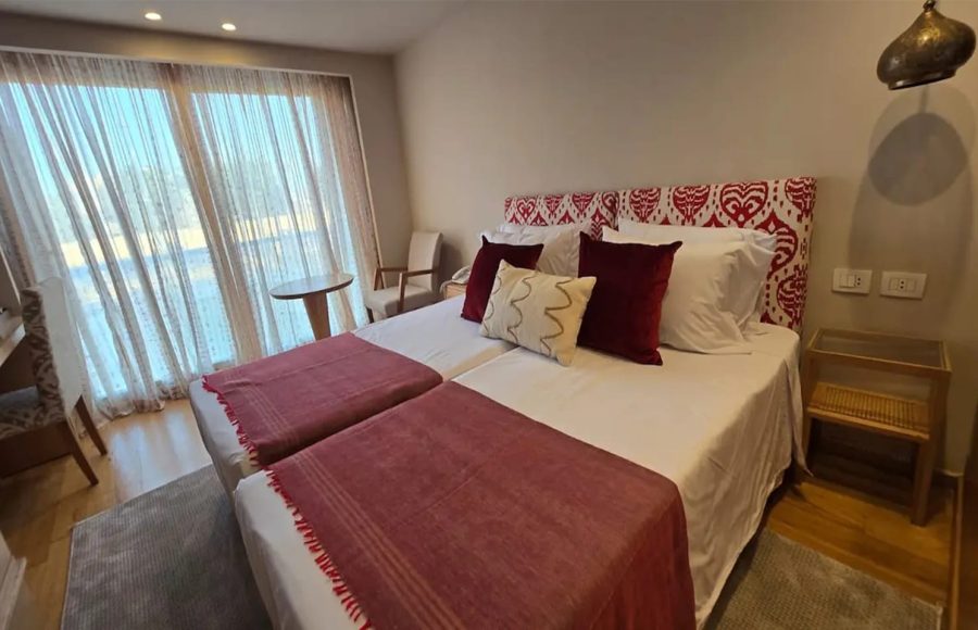 A cruise cabin with two twin beds featuring a red and white patterned headboard, a large window with sheer curtains, and wood flooring.