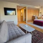 A cruise ship suite featuring a beige tufted sofa, flat-screen TV, and a large bed with a vibrant patterned headboard and rug.