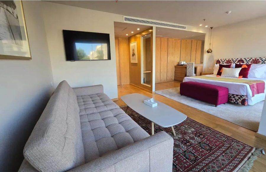 A cruise ship suite featuring a beige tufted sofa, flat-screen TV, and a large bed with a vibrant patterned headboard and rug.