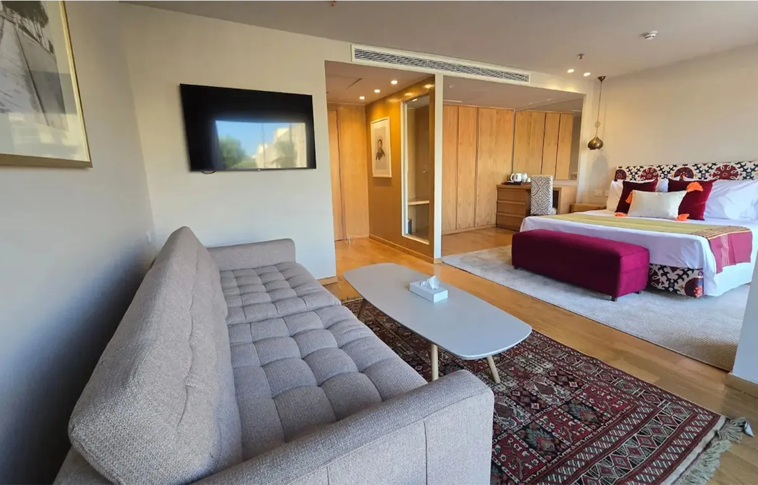 A cruise ship suite featuring a beige tufted sofa, flat-screen TV, and a large bed with a vibrant patterned headboard and rug.