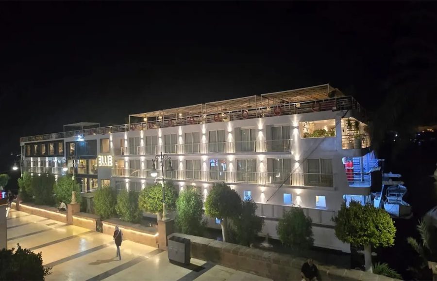 The MS Ellie Nile Cruise ship docked at night, brightly lit with exterior lighting against a dark sky and reflected on the boardwalk.