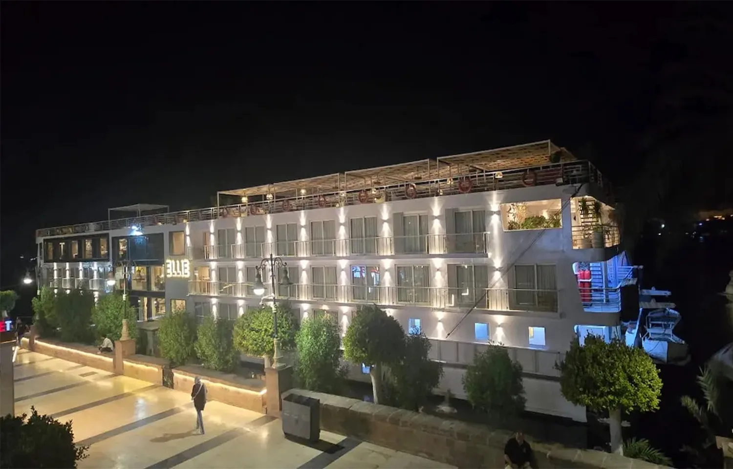 The MS Ellie Nile Cruise ship docked at night, brightly lit with exterior lighting against a dark sky and reflected on the boardwalk.