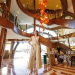 A guest standing in the grand lobby of the MS Esmeralda Nile Cruise, featuring a multi-level wooden staircase, striking hanging light fixture, and polished marble floors.
