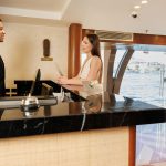 Friendly staff assisting a guest at the reception desk on the MS Esmeralda Nile Cruise, with scenic river views visible through the glass entrance.