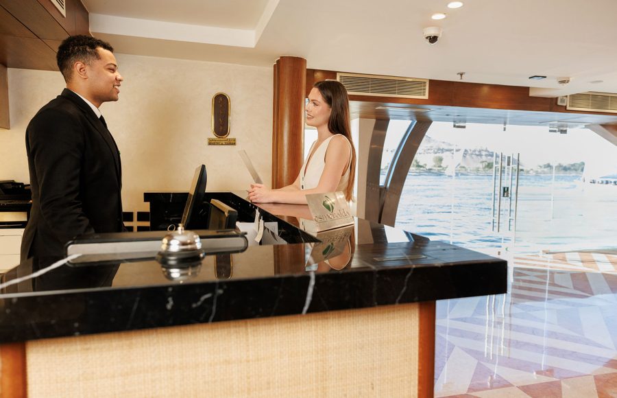 Friendly staff assisting a guest at the reception desk on the MS Esmeralda Nile Cruise, with scenic river views visible through the glass entrance.