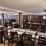 The interior dining room of the MS Hapi Nile cruise ship, featuring numerous dark wood tables and chairs set for service. The room has white ceilings with recessed lighting, wood-paneled pillars, and a decorative traditional lantern in the foreground.