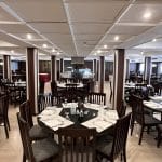 A wide interior view of the dining hall on the MS Hapi Nile cruise ship featuring rows of rectangular and round dark wood tables set for dinner under a white paneled ceiling with recessed lighting.