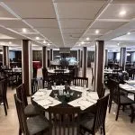 A wide interior view of the dining hall on the MS Hapi Nile cruise ship featuring rows of rectangular and round dark wood tables set for dinner under a white paneled ceiling with recessed lighting.