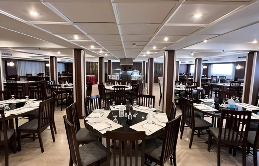 A wide interior view of the dining hall on the MS Hapi Nile cruise ship featuring rows of rectangular and round dark wood tables set for dinner under a white paneled ceiling with recessed lighting.