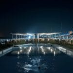 The sun deck of the MS Hapi cruise ship at night, featuring an illuminated swimming pool with a small fountain. The deck includes a seated bar or lounge area under a canopy with bright overhead lights.