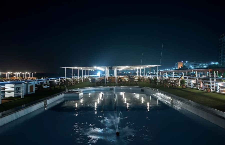 The sun deck of the MS Hapi cruise ship at night, featuring an illuminated swimming pool with a small fountain. The deck includes a seated bar or lounge area under a canopy with bright overhead lights.