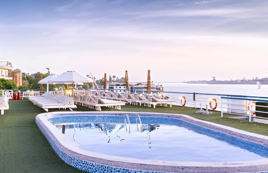 The sun deck of the MS Hapi Nile cruise ship featuring a small outdoor swimming pool. The deck is covered in green artificial turf and lined with white lounge chairs and sun umbrellas overlooking the river.