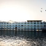A profile view of the MS Hapi Nile cruise ship on the water at sunset. Several birds are flying in the sky above the ship, and the bright sun is reflecting off the water.