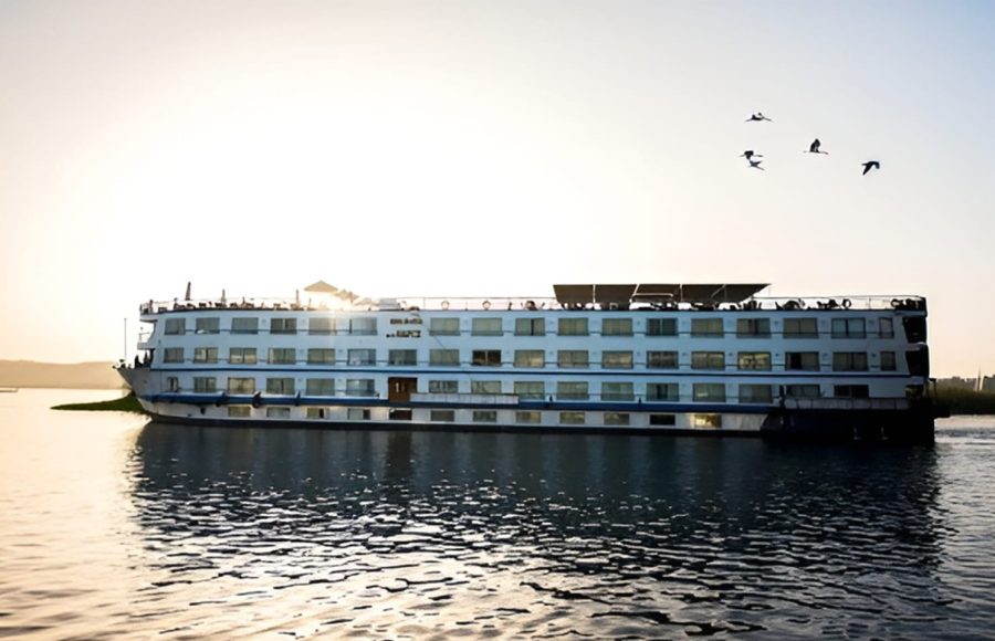 A profile view of the MS Hapi Nile cruise ship on the water at sunset. Several birds are flying in the sky above the ship, and the bright sun is reflecting off the water.