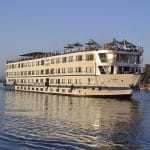 A large white multi-deck Nile cruise ship named MS Hapi sailing on the Nile River during the day. The ship features several levels of guest cabins with windows, a sun deck with beige umbrellas at the top, and a traditional felucca sailboat visible in the background against a clear blue sky.