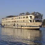 A large white multi-deck Nile cruise ship named MS Hapi sailing on the Nile River during the day. The ship features several levels of guest cabins with windows, a sun deck with beige umbrellas at the top, and a traditional felucca sailboat visible in the background against a clear blue sky.
