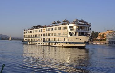 A large white multi-deck Nile cruise ship named MS Hapi sailing on the Nile River during the day. The ship features several levels of guest cabins with windows, a sun deck with beige umbrellas at the top, and a traditional felucca sailboat visible in the background against a clear blue sky.