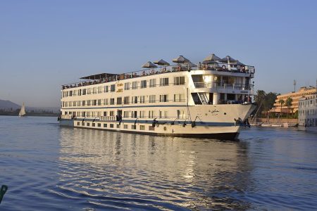 A Large White Multi-Deck Nile Cruise Ship Named Ms Hapi Sailing On The Nile River During The Day. The Ship Features Several Levels Of Guest Cabins With Windows, A Sun Deck With Beige Umbrellas At The Top, And A Traditional Felucca Sailboat Visible In The Background Against A Clear Blue Sky.