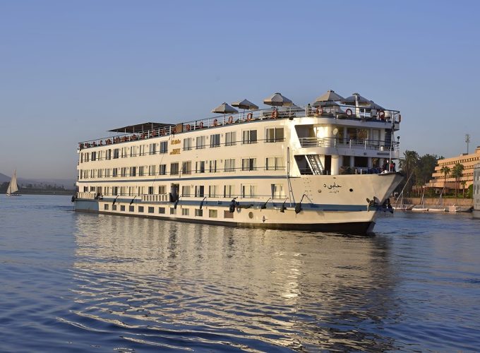 A Large White Multi-Deck Nile Cruise Ship Named Ms Hapi Sailing On The Nile River During The Day. The Ship Features Several Levels Of Guest Cabins With Windows, A Sun Deck With Beige Umbrellas At The Top, And A Traditional Felucca Sailboat Visible In The Background Against A Clear Blue Sky.