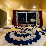 A hotel room cabin on the MS Hapi Nile Cruise featuring a twin bed decorated with an elaborate blue and white striped towel animal in the shape of a swan. The room has warm yellow lighting, a large window with red curtains, a television, and a small sitting area.