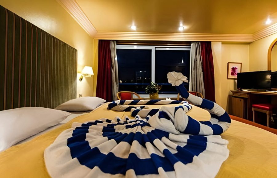 A hotel room cabin on the MS Hapi Nile Cruise featuring a twin bed decorated with an elaborate blue and white striped towel animal in the shape of a swan. The room has warm yellow lighting, a large window with red curtains, a television, and a small sitting area.