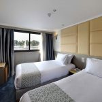A cozy and well-appointed Cabin Twin Bed on the MS Nile Treasure Cruise, featuring two single beds, crisp white linens, a cushioned headboard, and a large window overlooking the river.
