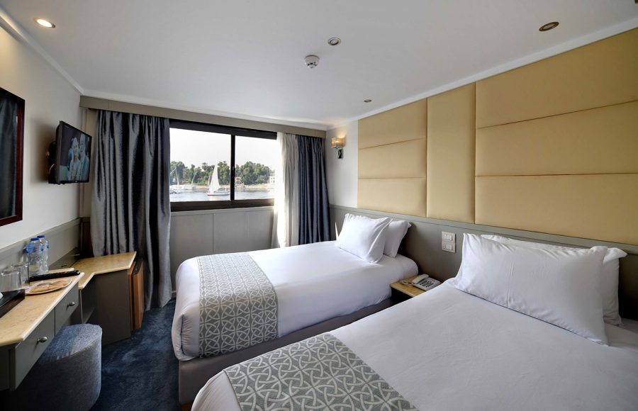 A cozy and well-appointed Cabin Twin Bed on the MS Nile Treasure Cruise, featuring two single beds, crisp white linens, a cushioned headboard, and a large window overlooking the river.