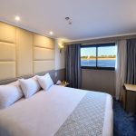 A deluxe suite aboard the MS Nile Treasure Cruise, featuring a plush king-size bed, upholstered headboard, vanity area, and a large window framing scenic Nile views.