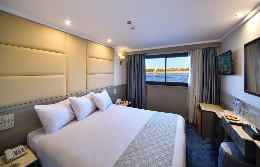 A deluxe suite aboard the MS Nile Treasure Cruise, featuring a plush king-size bed, upholstered headboard, vanity area, and a large window framing scenic Nile views.