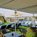 A shaded sun deck restaurant area on the MS Nile Treasure Cruise, featuring outdoor dining tables, blue-and-white striped loungers, and panoramic views of the Nile River banks.