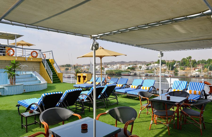 A shaded sun deck restaurant area on the MS Nile Treasure Cruise, featuring outdoor dining tables, blue-and-white striped loungers, and panoramic views of the Nile River banks.