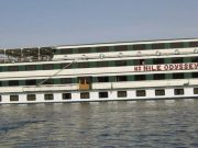 The exterior of a multi-deck white and green cruise ship named "MS NILE ODYSSEY" floating on the river. The name is written in large red letters on the middle deck.