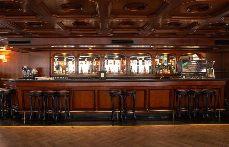 Elegant cruise ship bar with a long, polished wood counter, tiered shelves of illuminated liquor bottles, and a row of dark leather bar stools.