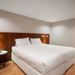 Modern cruise ship cabin with a large bed, polished wood headboard, a white nightstand, and a full-length mirror.