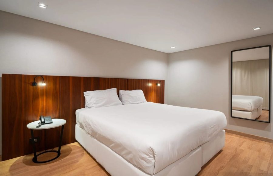 Modern cruise ship cabin with a large bed, polished wood headboard, a white nightstand, and a full-length mirror.