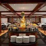 Buffet service in the elegant MS Nile Plaza dining room, showing a central food display, stacked plates, and surrounding dining tables.