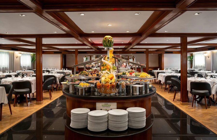 Buffet service in the elegant MS Nile Plaza dining room, showing a central food display, stacked plates, and surrounding dining tables.