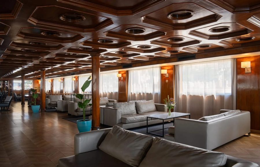 Long, luxurious lounge on the MS Nile Plaza with a high-gloss octagonal coffered wood ceiling, multiple gray leather seating groups, and large windows.