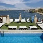 Swimming pool and sun loungers on the MS Nile Plaza sun deck, with a panoramic view of the wide Nile River and the city shoreline.