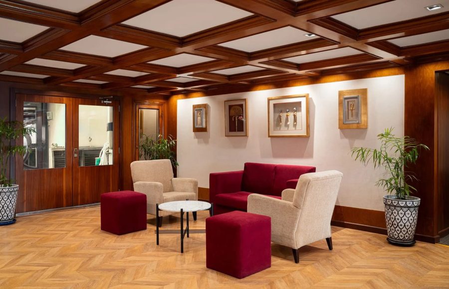 Cozy lounge area on the MS Nile Plaza with beige armchairs, red ottomans, and a coffered wood ceiling, featuring Egyptian-themed artwork.